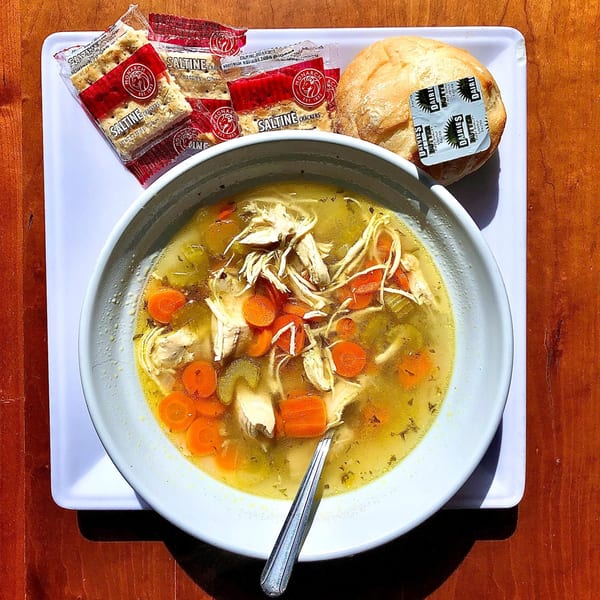 A bowl of Knickerbockers Deli chicken noodle soup with carrots and celery, served with crackers and a roll.