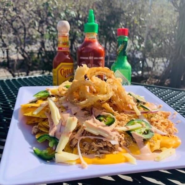 Knickerbockers Deli nachos with cheese, meat, jalapeños, and onion rings, with hot sauce bottles in the background.