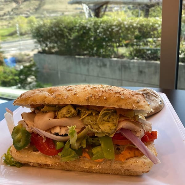 A Knickerbockers Deli sandwich with turkey, lettuce, tomatoes, onions, and artichokes on a white plate.