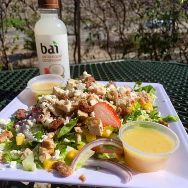 A vibrant salad with chicken, strawberries, feta, and dressing, alongside a Bai drink, on an outdoor table.